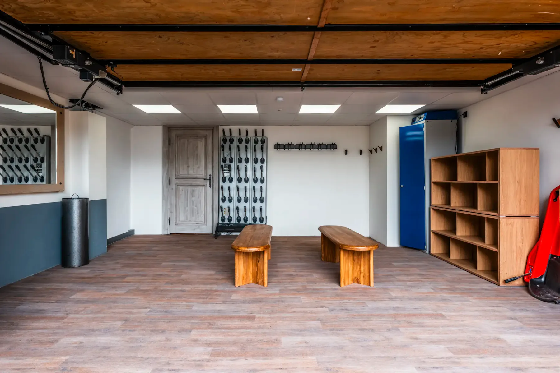Spacious room with wooden benches, wall hooks, a mirror, shelving, and a blue door. Ceiling lights illuminate the wooden floor.