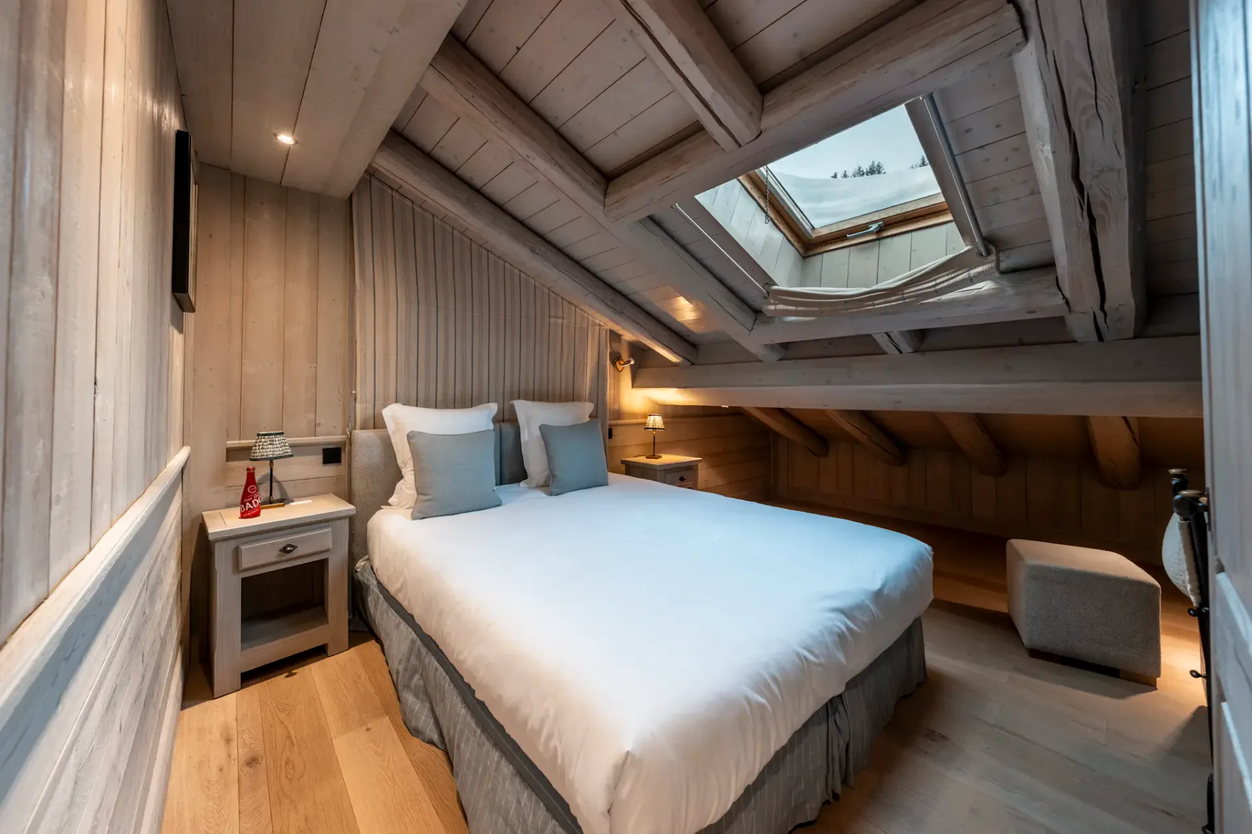 Cozy attic bedroom with wooden walls, a skylight, a neatly made bed, side tables, and soft lighting, creating a warm and inviting atmosphere.