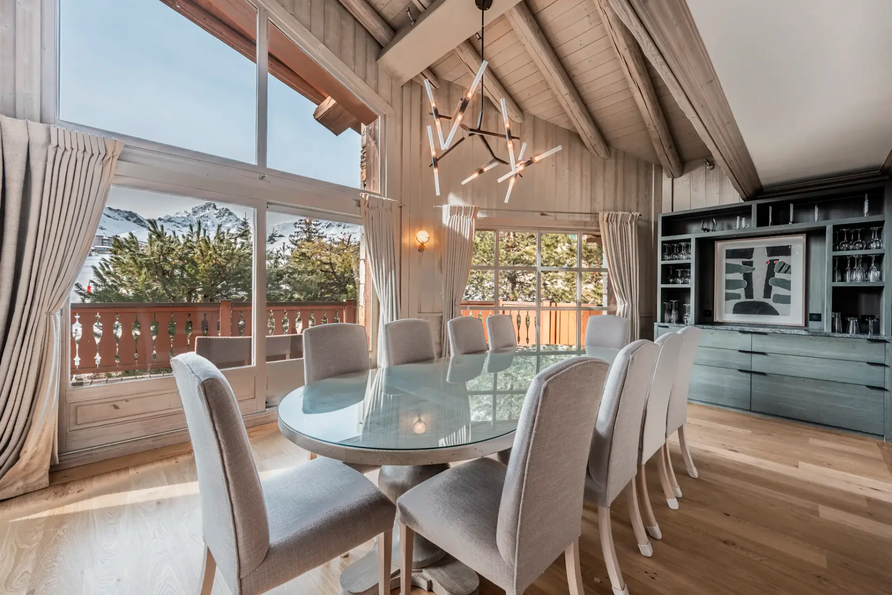 A dining room with a glass table, ten upholstered chairs, a modern chandelier, large windows, and mountain views.