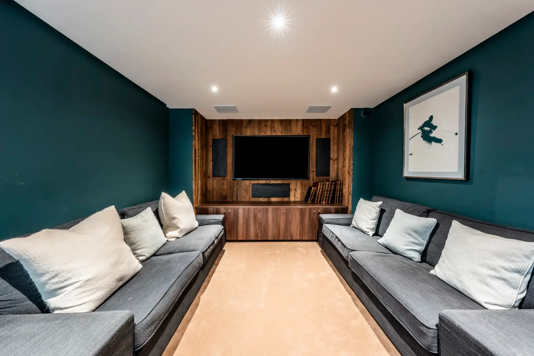 Modern home theater with dark teal walls, two gray sofas, a wall-mounted TV on wooden paneling, and a framed picture.