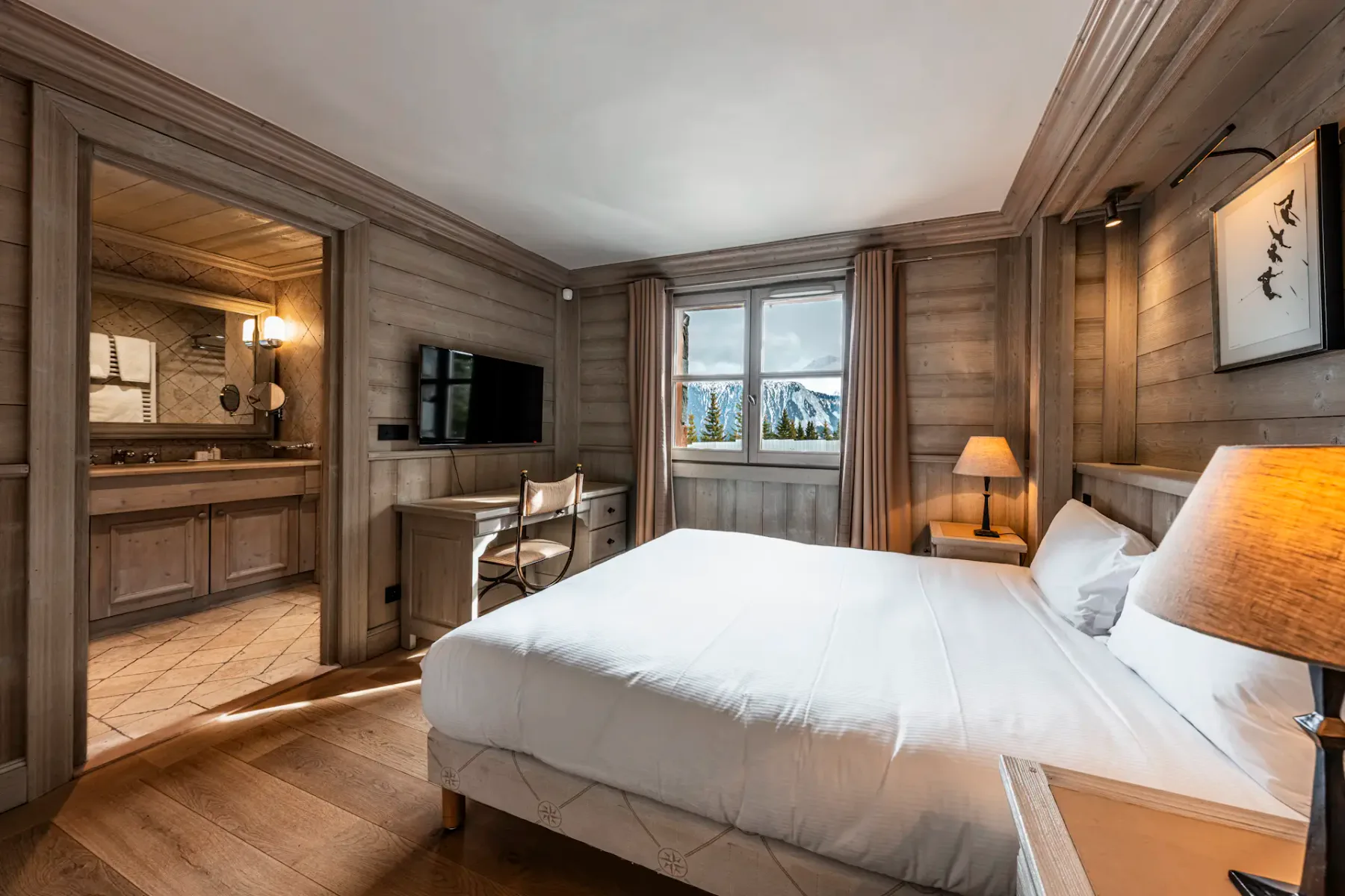 Cozy hotel room with a large bed, wooden walls, a desk, and a window view of snowy mountains. En-suite bathroom visible to the left.