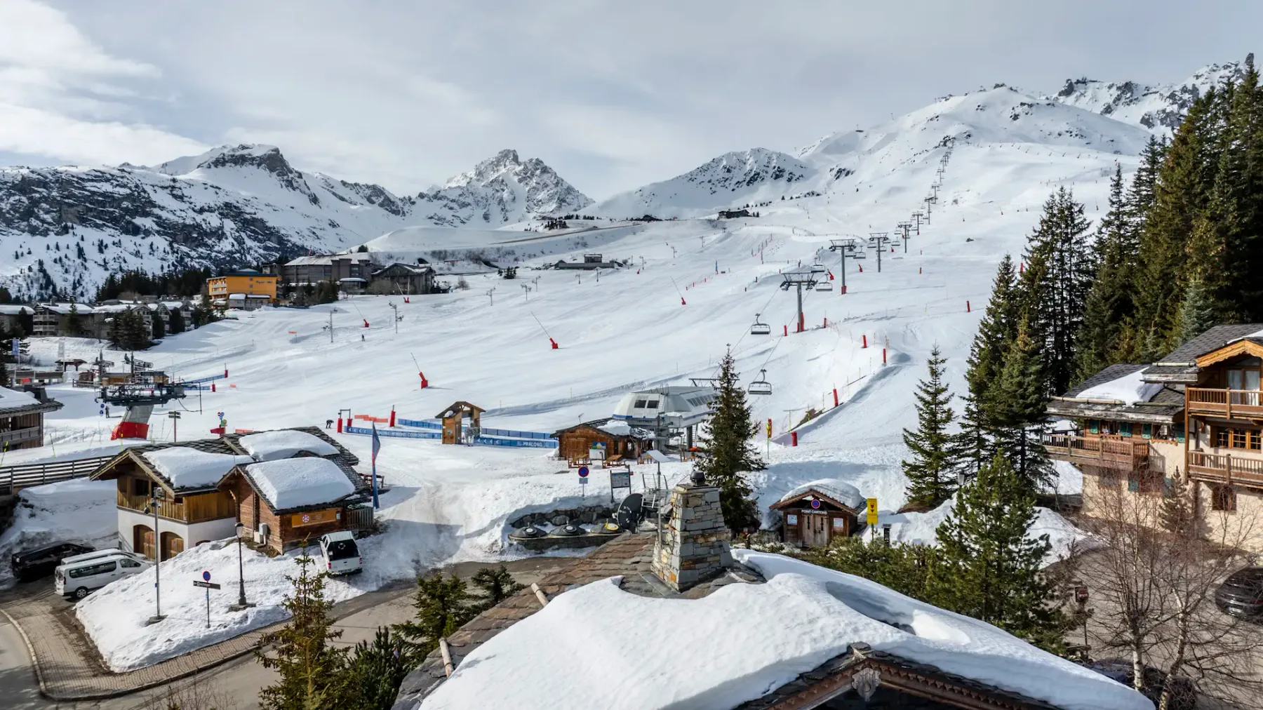 Snow-covered ski resort with chalet-style buildings, ski lifts, and mountainous backdrop under a cloudy sky. Pine trees line the foreground.