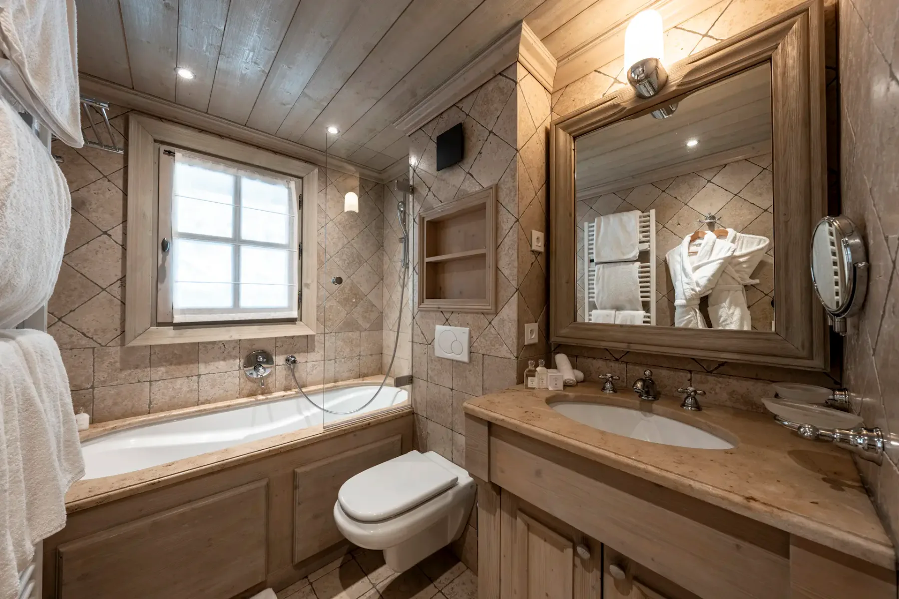 Cozy bathroom with wooden accents, featuring a bathtub, toilet, double sink, mirror, and window. Tiled walls and ceiling lights add warmth.