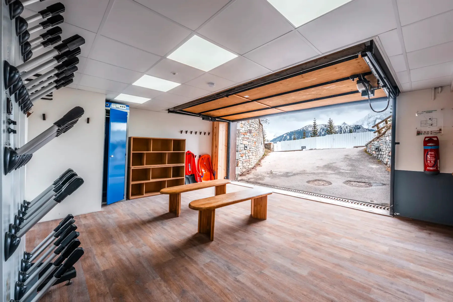 Spacious ski storage room with wooden benches, racks for gear, and an open garage door revealing a snowy mountain landscape outside.