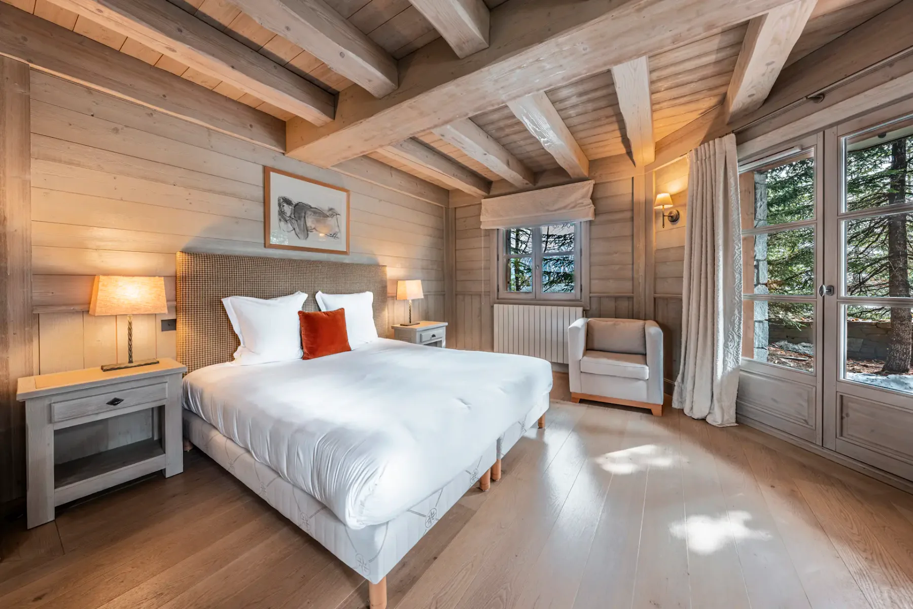 Cozy bedroom with wooden walls, a large bed, white bedding, a red accent pillow, bedside lamps, an armchair, and a window with forest view.