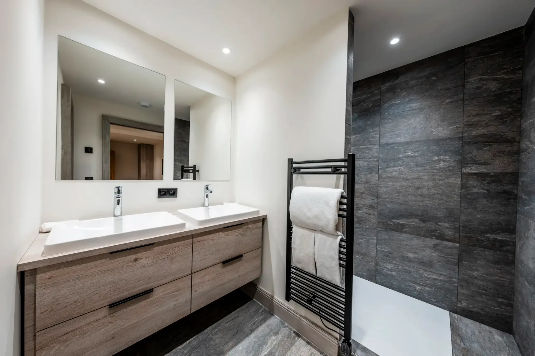 Modern bathroom with dual sinks, large mirrors, a dark tiled shower, and a towel rack. Neutral tones and sleek design create a clean look.