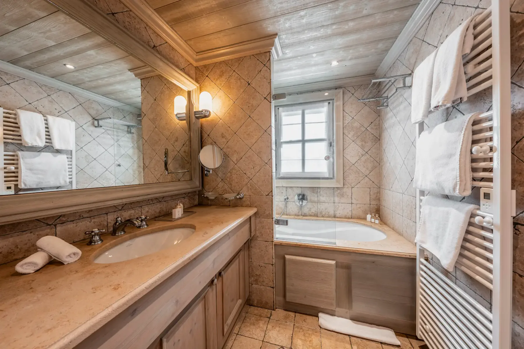 Rustic bathroom with wooden ceiling, large mirror, dual sinks, and a bathtub. Tiled walls and heated towel rack with folded white towels.