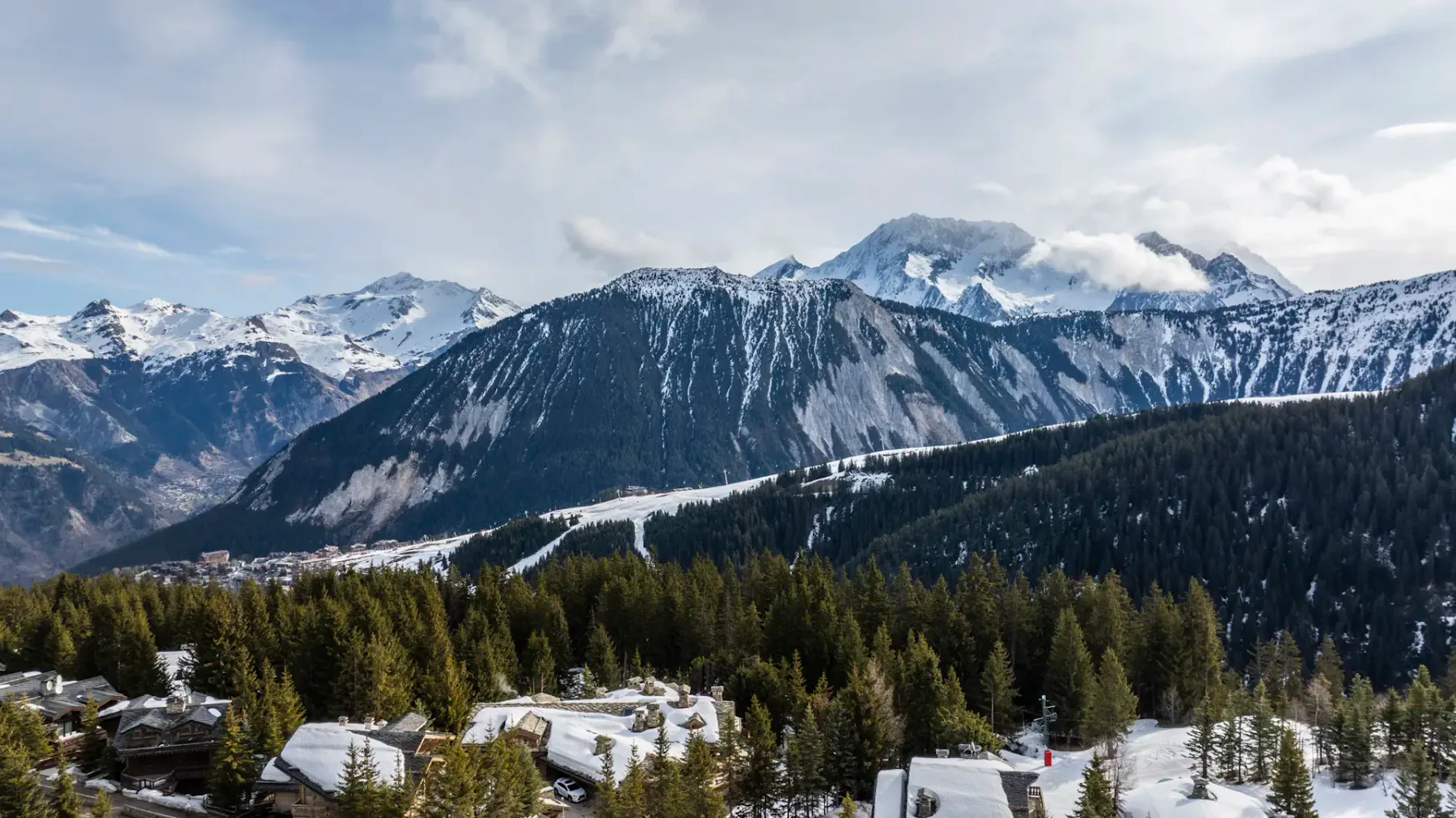 Snow-covered mountains and dense evergreen forests under a cloudy sky, with rooftops of buildings visible in the foreground.