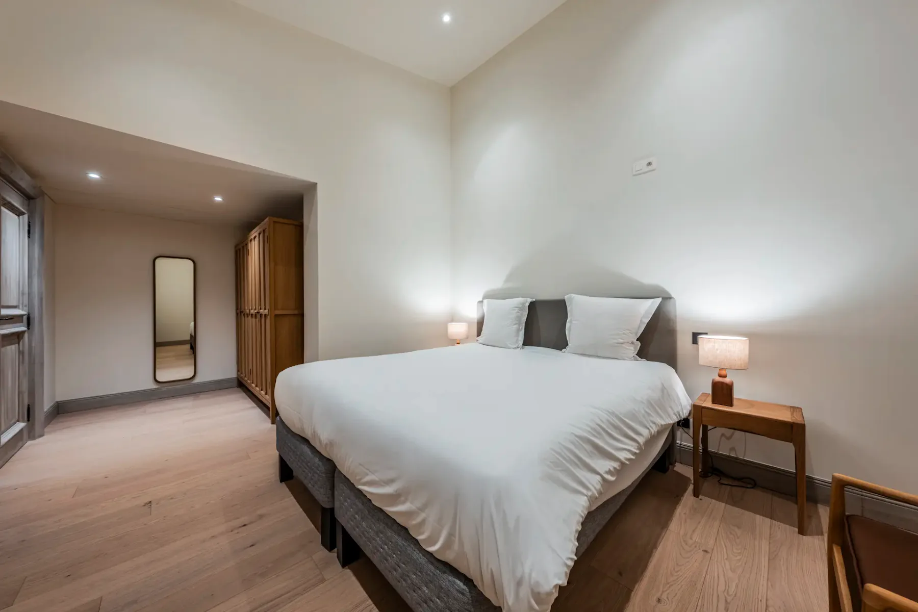 Minimalist bedroom with a large bed, white bedding, bedside lamps, wooden wardrobe, and a tall mirror on a light wooden floor.