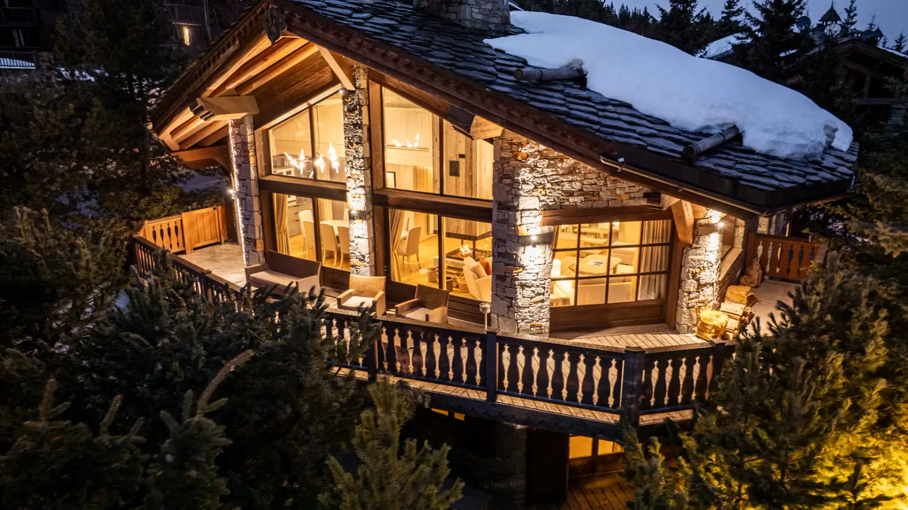 Cozy chalet with large windows, warm interior lighting, and snow-covered roof, surrounded by pine trees at dusk.