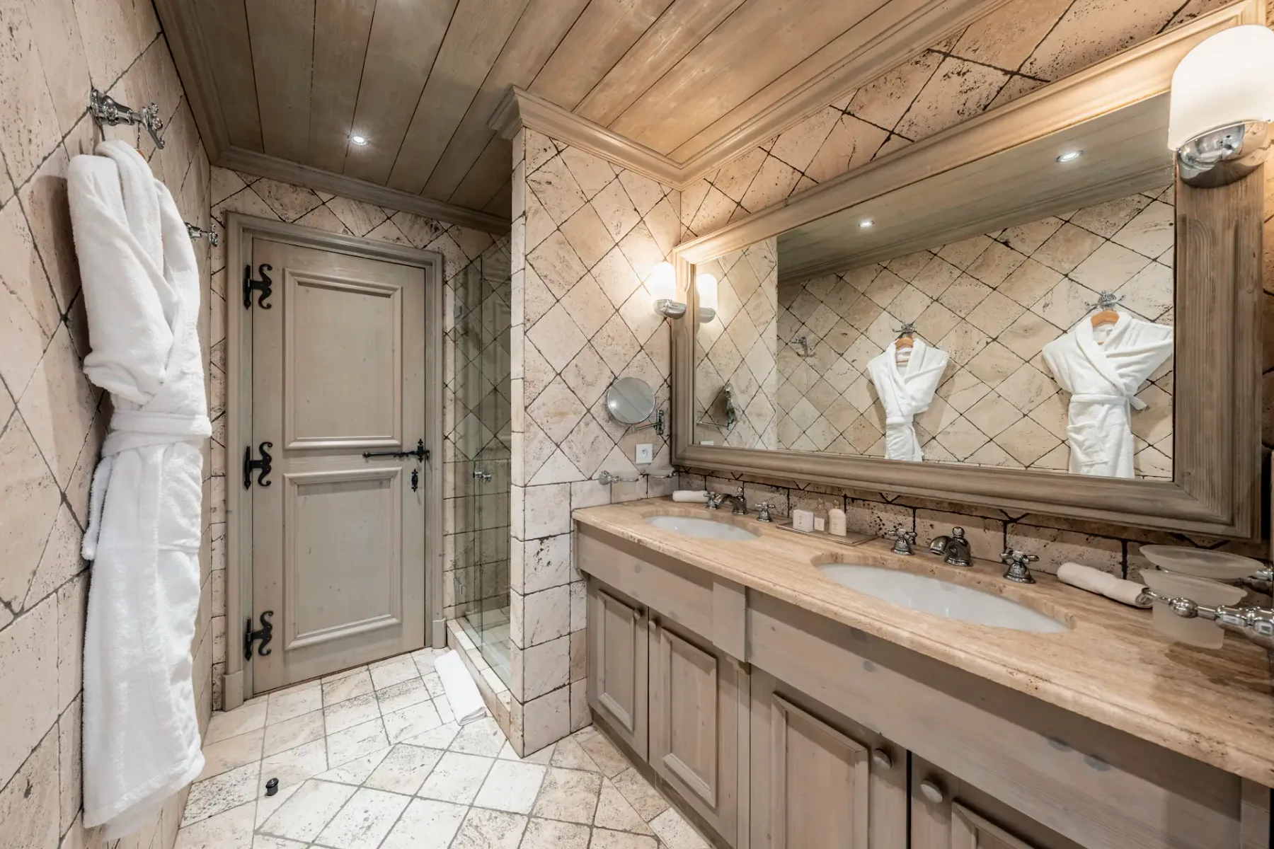 Luxurious bathroom with a large mirror, double sink, tiled walls, wooden ceiling, and a hanging white robe beside a closed door.
