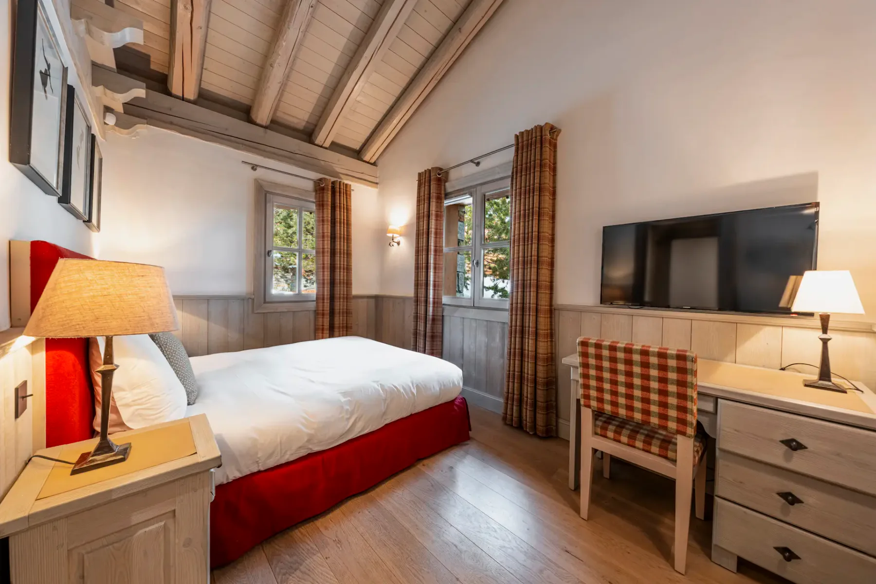 Cozy bedroom with a double bed, plaid curtains, wooden ceiling, desk with a lamp, and a wall-mounted TV. Warm lighting creates a welcoming atmosphere.