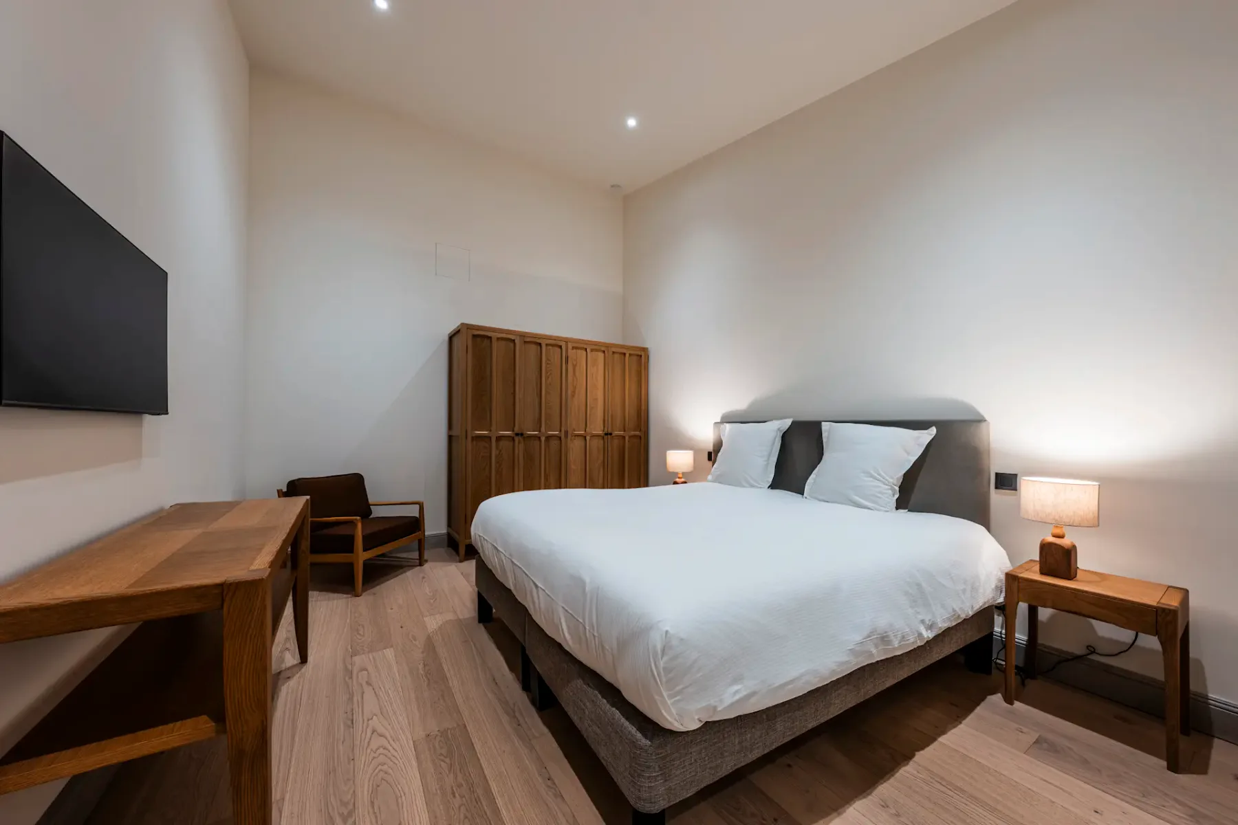 Minimalist bedroom with a double bed, white bedding, wooden furniture, a wall-mounted TV, and soft lighting from bedside lamps.