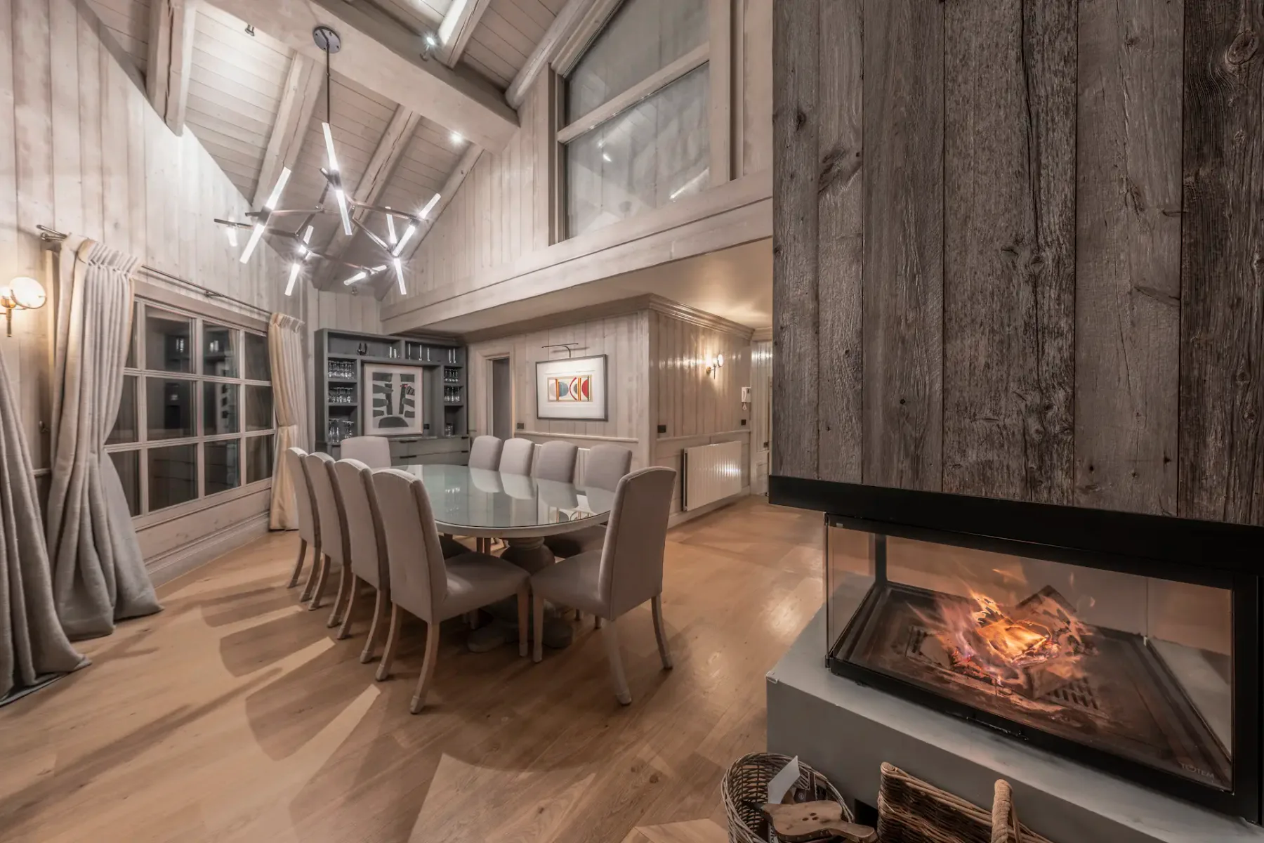 Cozy dining room with a large table, modern chandelier, and a lit fireplace. Wooden walls and floor create a warm, inviting atmosphere.
