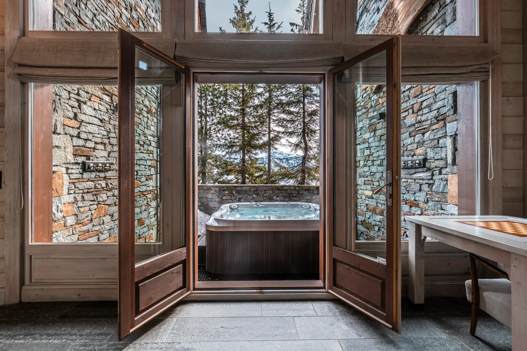 Open wooden doors lead to an outdoor hot tub surrounded by stone walls, with a view of trees and mountains in the background.