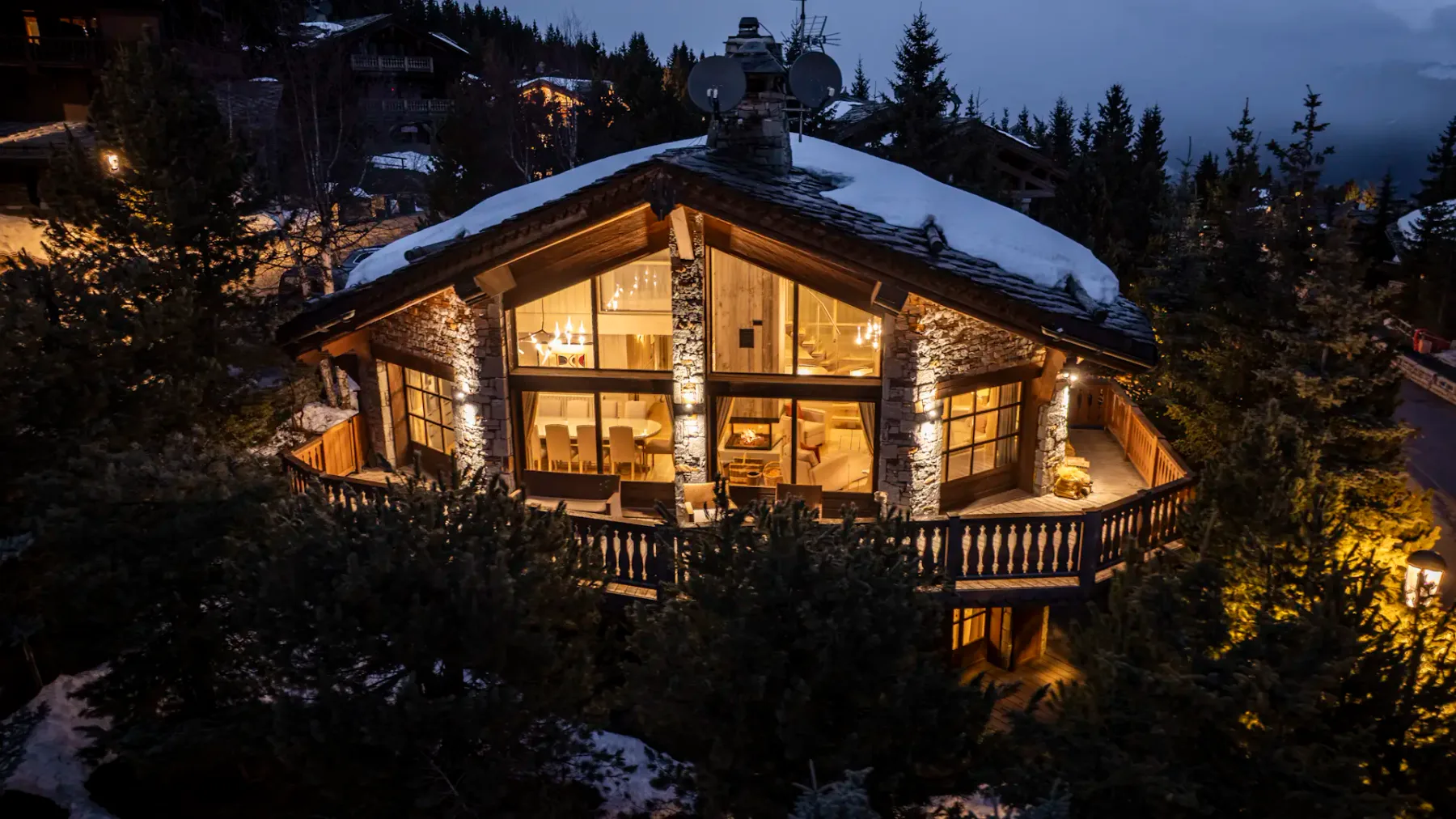 A cozy, illuminated chalet with large windows, surrounded by snow-dusted trees, set in a mountainous, twilight landscape.