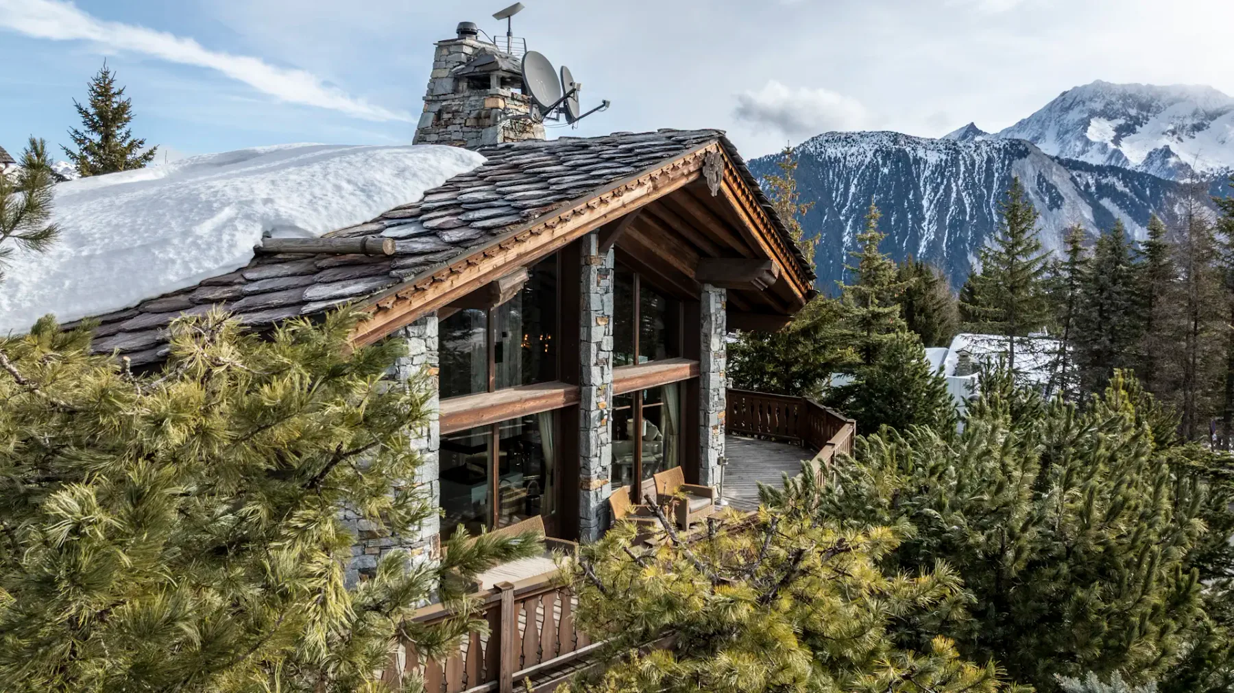 A rustic mountain cabin with a stone chimney, surrounded by snow-covered trees, set against a backdrop of snowy peaks and a clear sky.