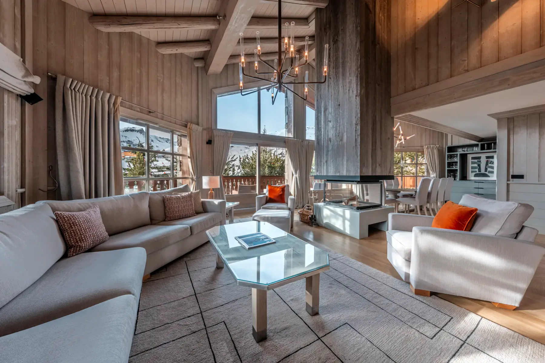 Spacious living room with large windows, white sofas, orange pillows, a glass coffee table, and a chandelier, set in a rustic wooden interior.