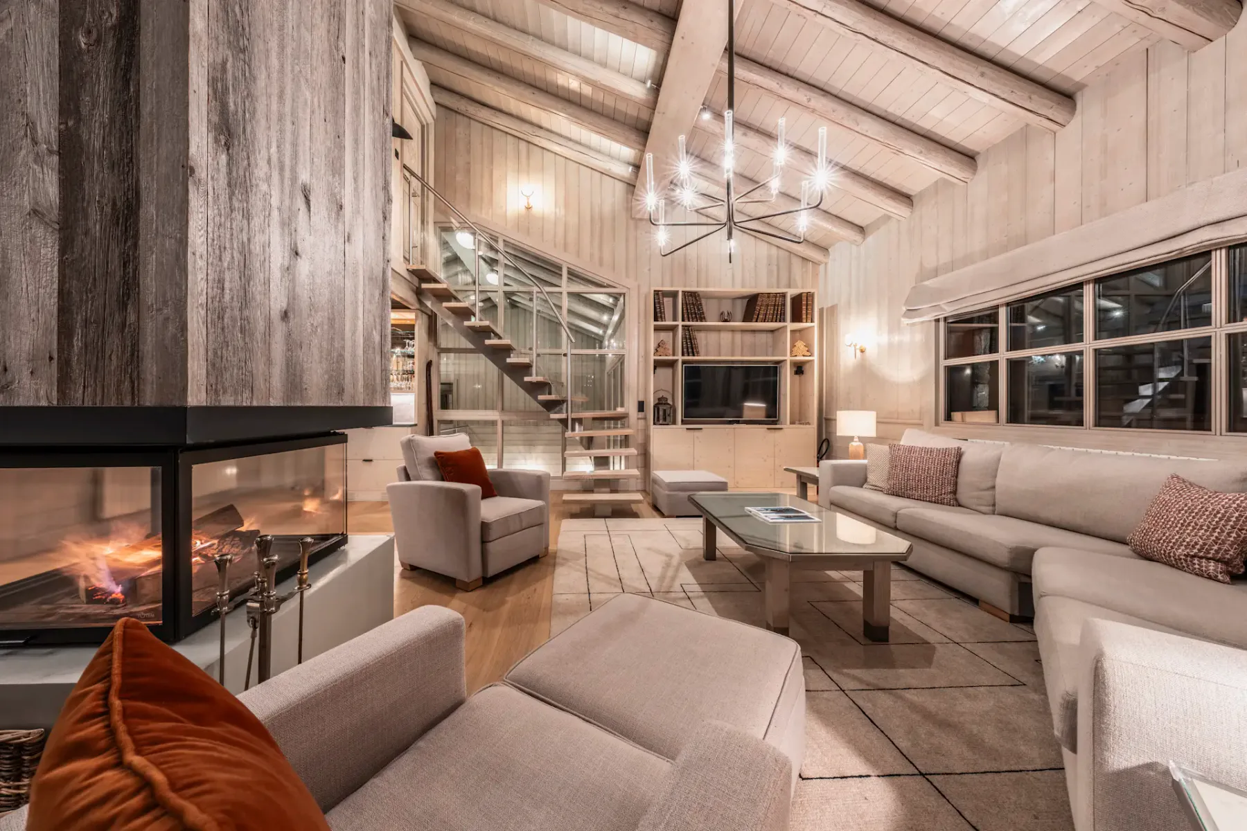 Cozy living room with beige sofas, a modern fireplace, a glass coffee table, and a chandelier. Wooden walls and ceiling with exposed beams.