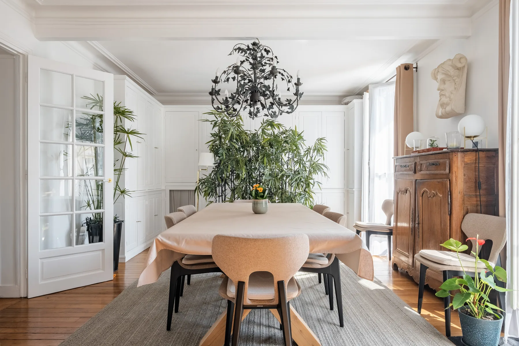A bright dining room with a long table, chairs, and leafy plants.