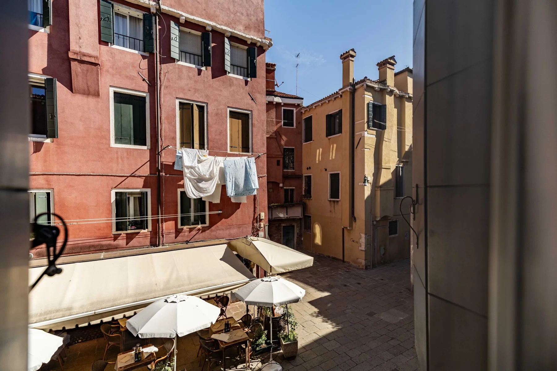 Un patio soleado entre edificios coloridos, con mesas de café bajo toldos.