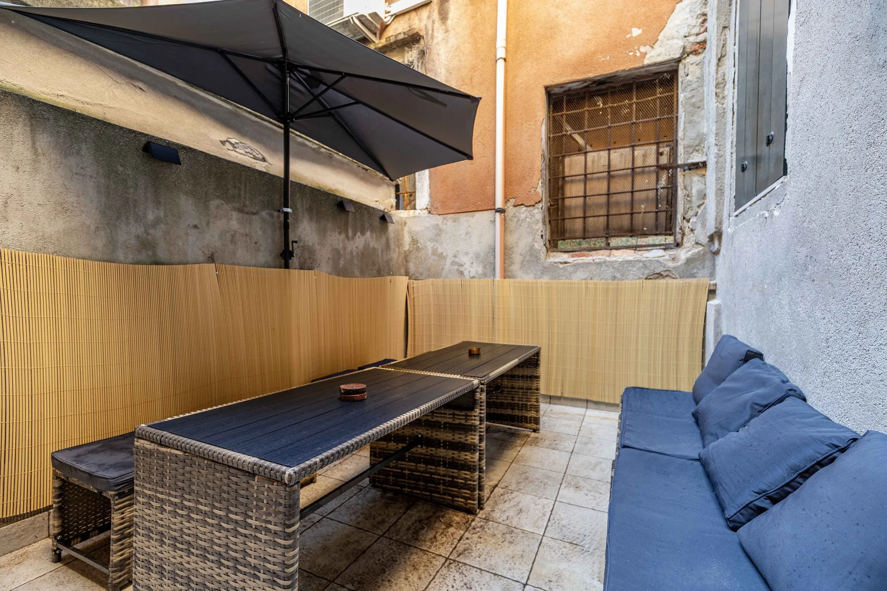 Un patio exterior con una mesa larga y asientos azules bajo una sombrilla grande.