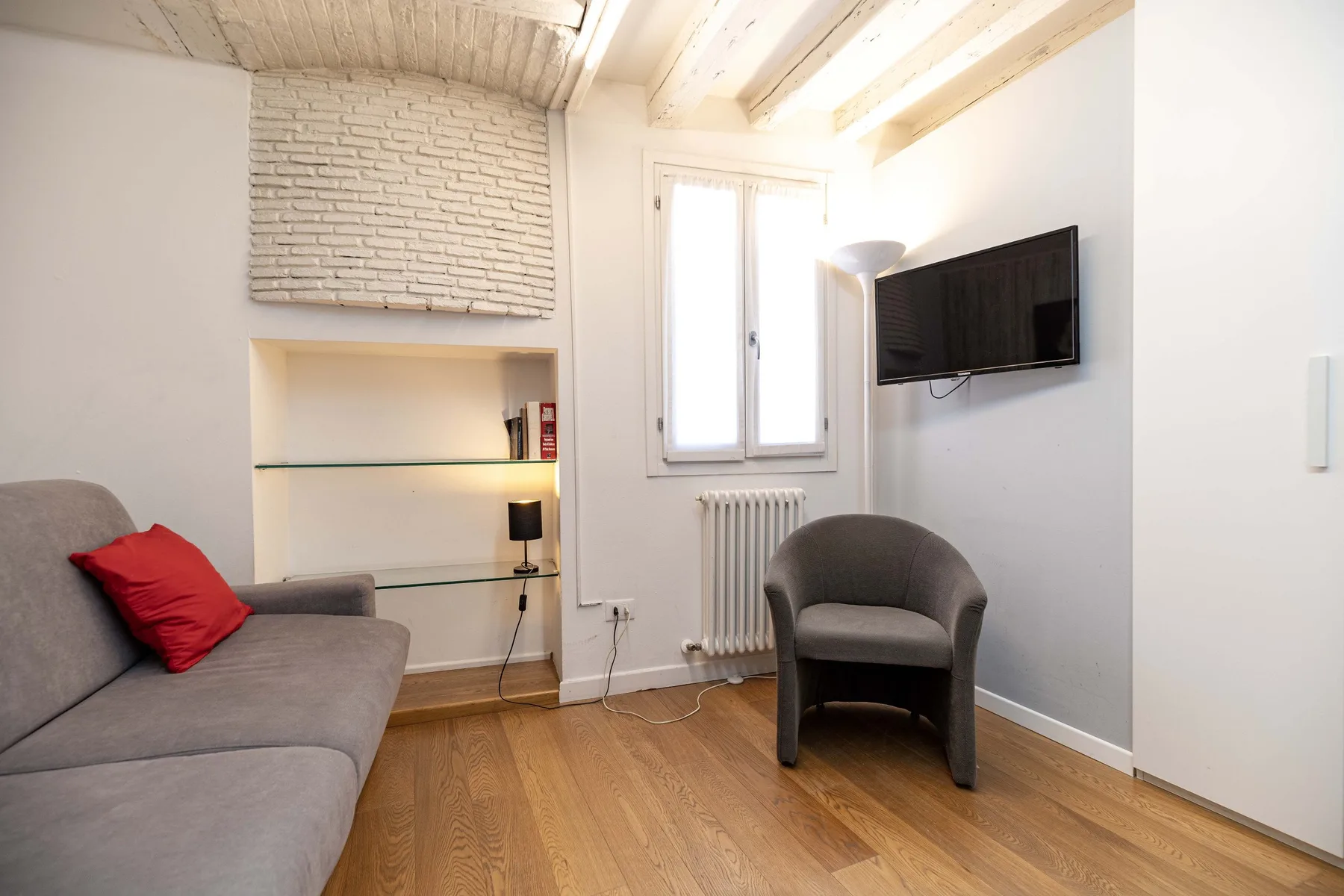 Una sala luminosa con un sofá gris, un sillón y un televisor montado en la pared.
