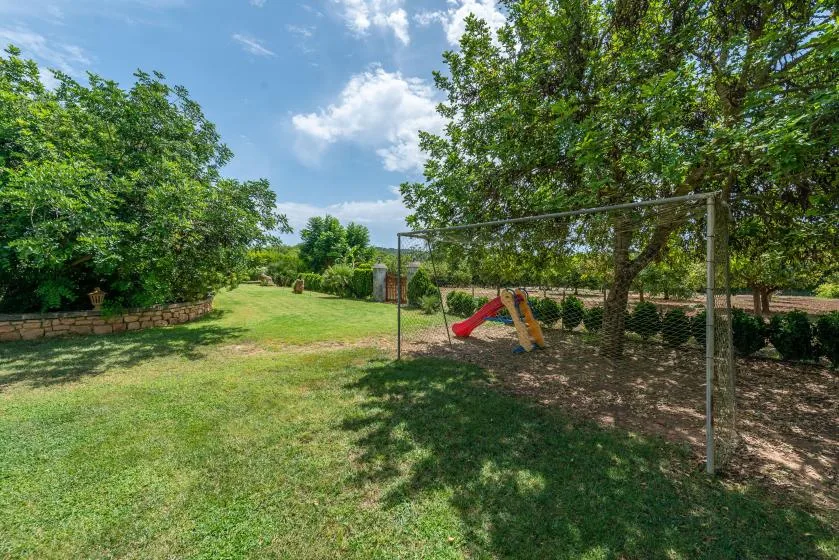 Un jardín con césped y un pequeño tobogán infantil bajo árboles frondosos.