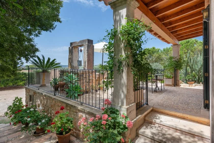 Un patio cubierto con barandilla negra, flores en macetas y vistas al jardín.