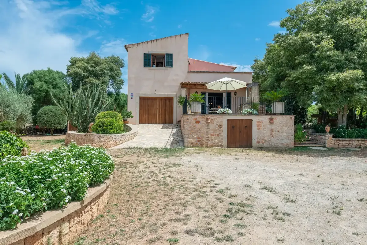 Una casa de dos plantas se alza tras un patio seco, rodeada de árboles y plantas con flores.