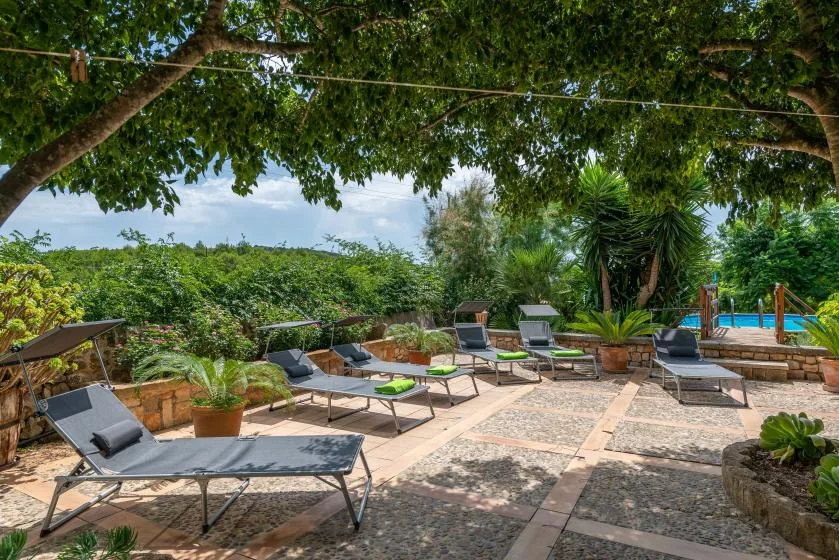An outdoor patio with lounge seating sits beneath leafy trees beside a swimming pool.
