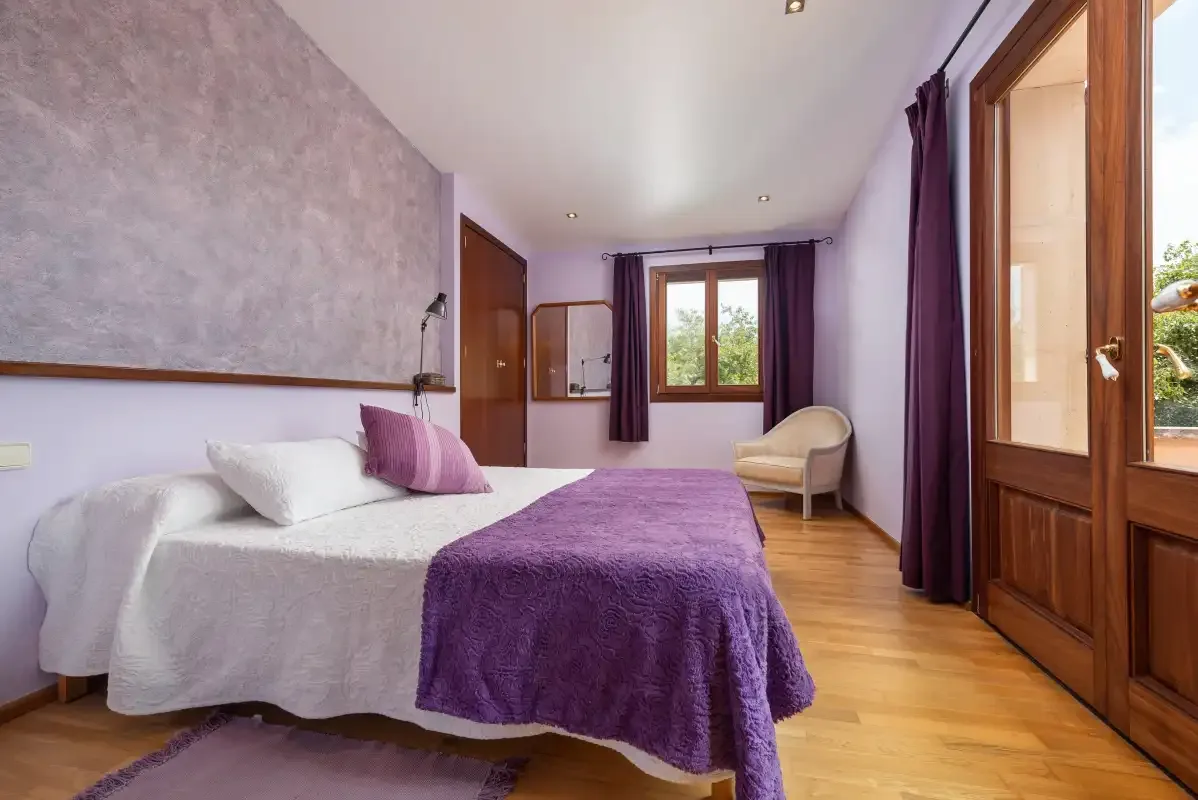 Un dormitorio luminoso con una cama vestida de morado y una ventana con vistas a la vegetación.