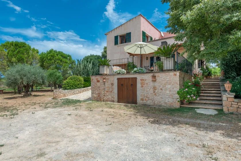 Una casa de dos plantas se alza sobre un muro de piedra, con patio y escaleras exteriores.