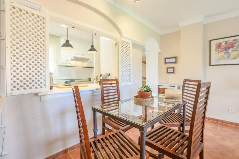 A dining area with a glass-top table and wooden seating beside an open kitchen.