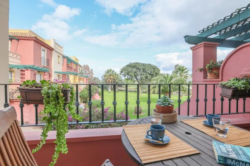 A terrace dining area overlooks a garden with palm trees and pink buildings.