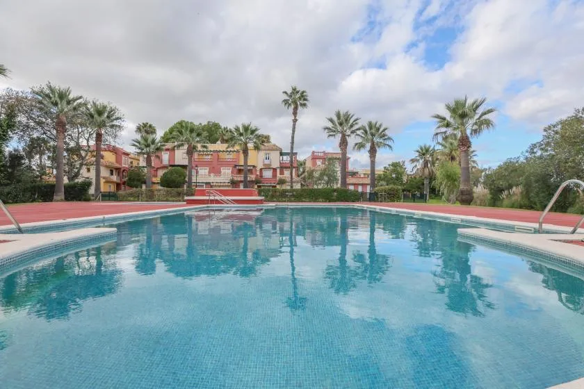A swimming pool reflects palm trees and colorful buildings under a partly cloudy sky.