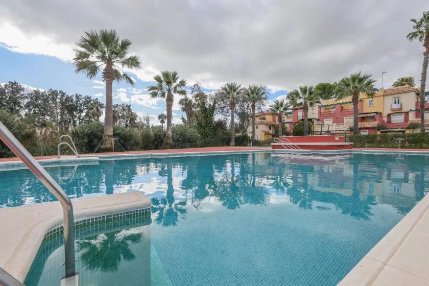 An outdoor swimming pool reflects palm trees and colorful buildings under a cloudy sky.