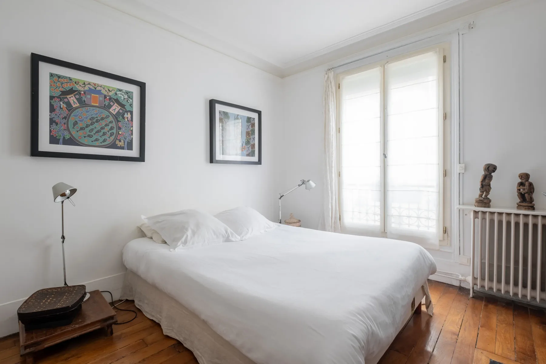 A bright bedroom with a white bed, framed artwork, and a large window.