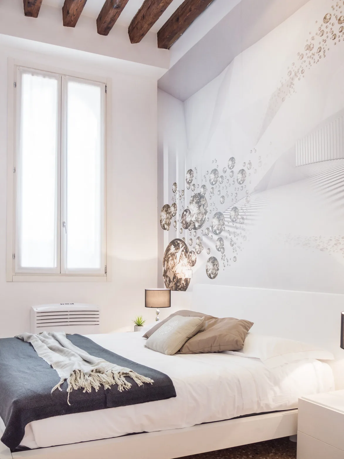 Un dormitorio luminoso con cama, lámpara de noche y ventana alta.
