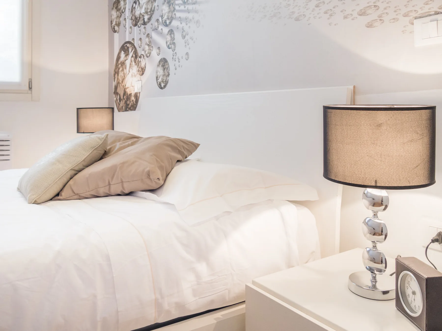 Un dormitorio luminoso con ropa de cama blanca, almohadas neutras y lámparas de noche.