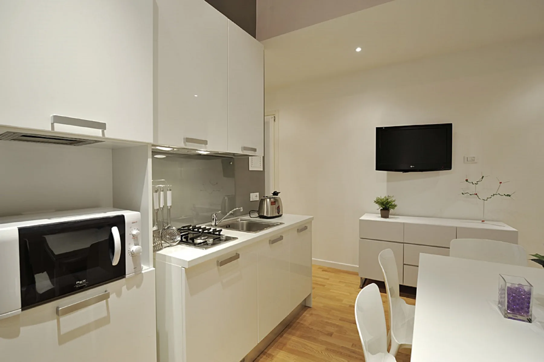 Una cocina compacta y zona de comedor con armarios blancos y un televisor en la pared.
