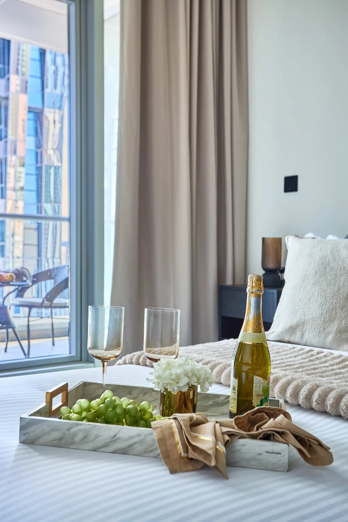 Un dormitorio con una cama y una bandeja de uvas, flores y una botella junto a la ventana.