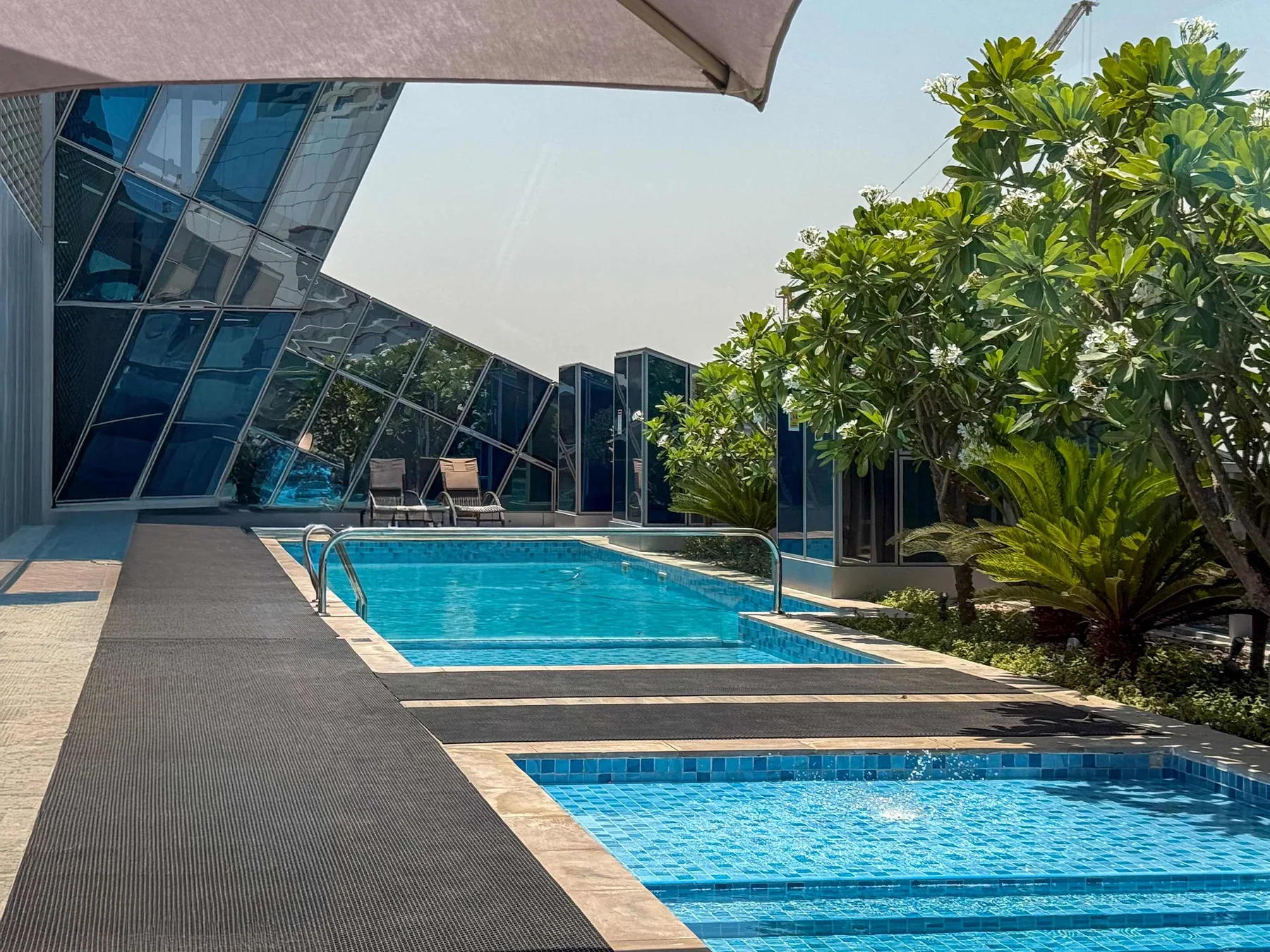 Una zona de piscina al aire libre con agua azul, vegetación y un edificio moderno de vidrio.