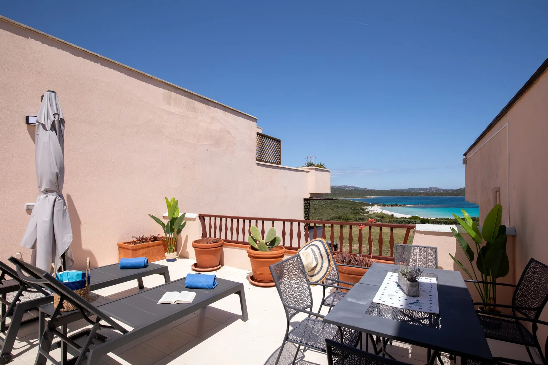 A sunny terrace with outdoor seating, potted plants, and a view of the sea.