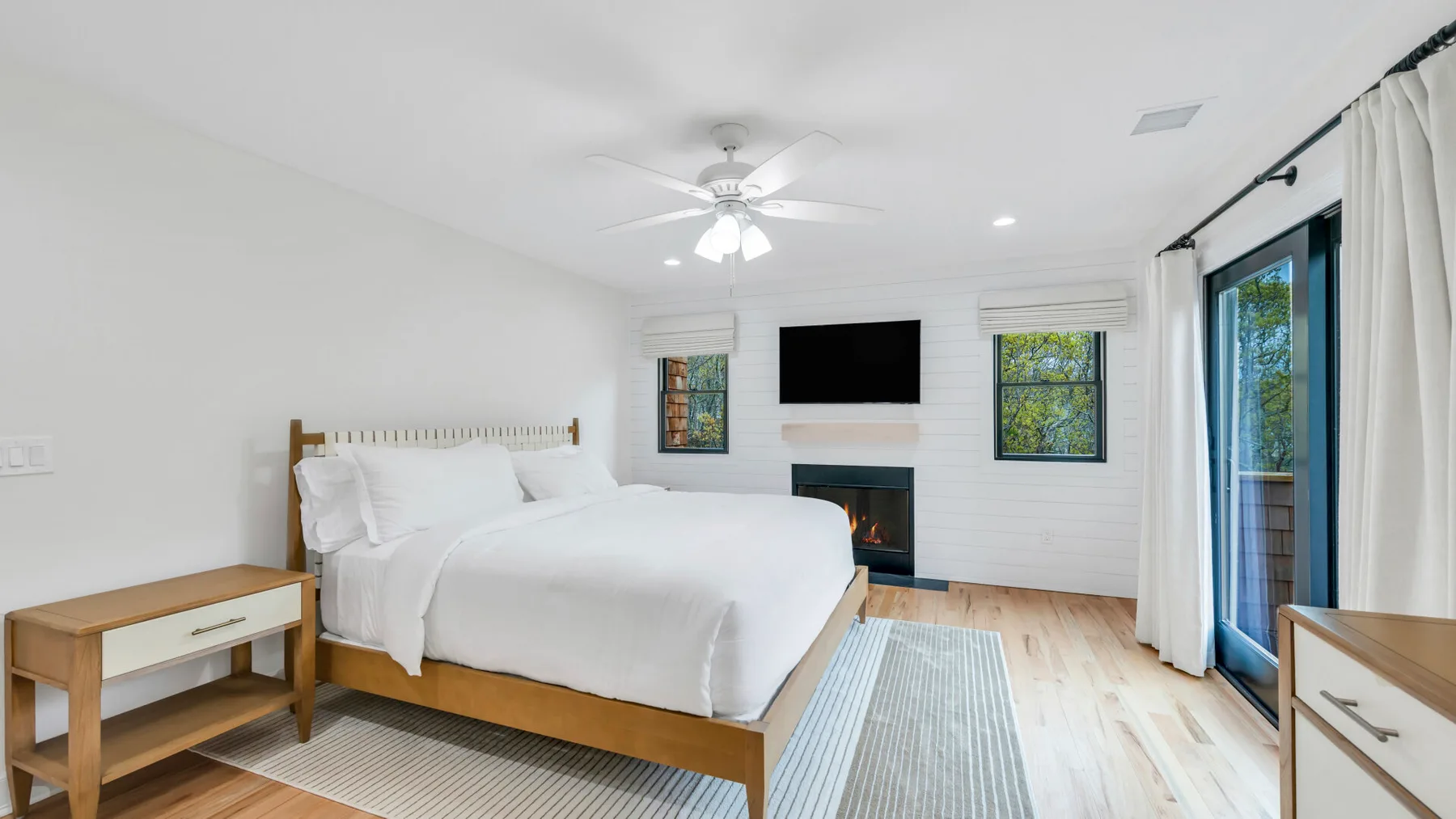 Un dormitorio luminoso con una cama bien tendida, un televisor en la pared y ventanas grandes.