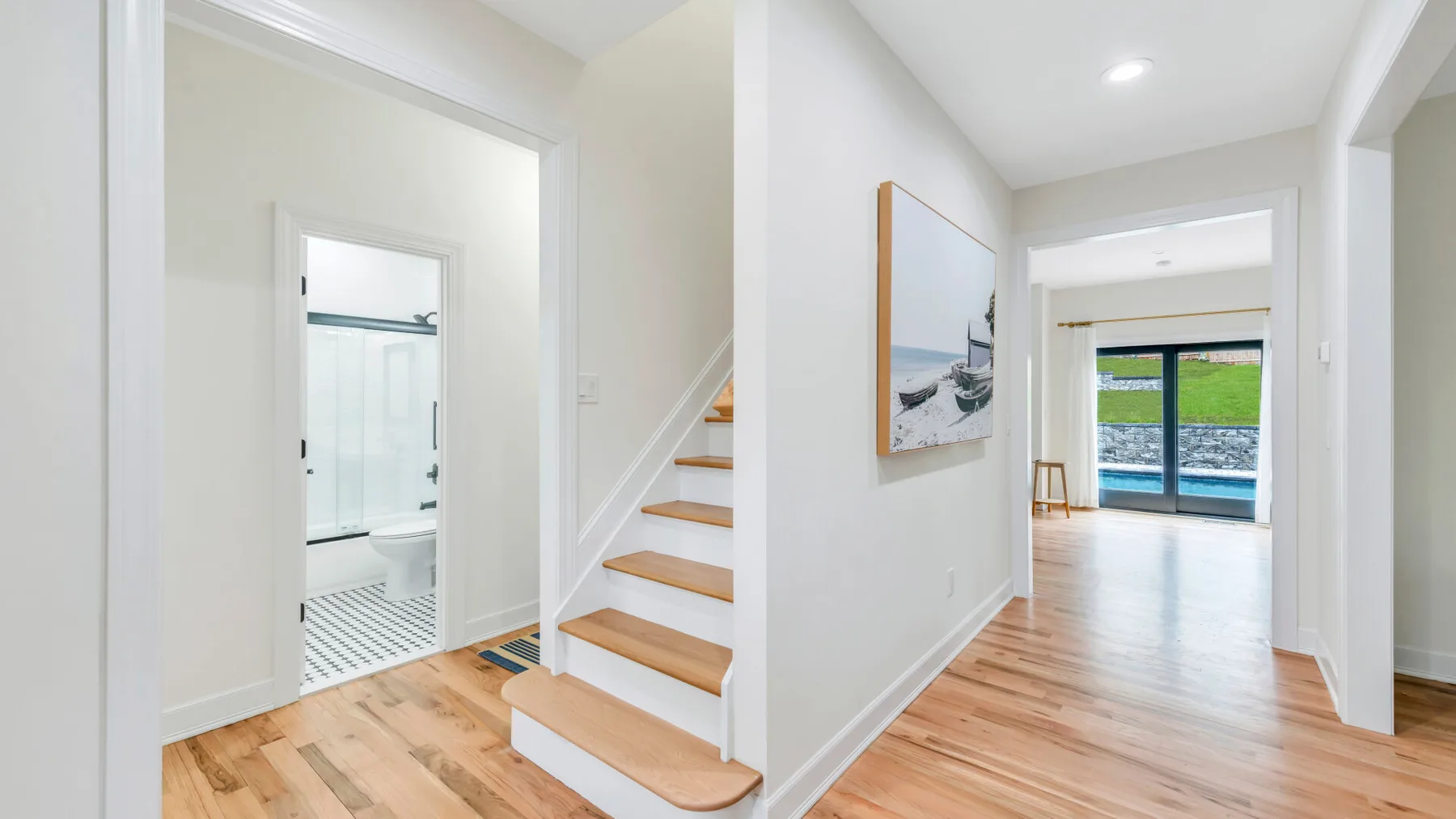 Un pasillo luminoso conecta un baño y una escalera, con suelo de madera.