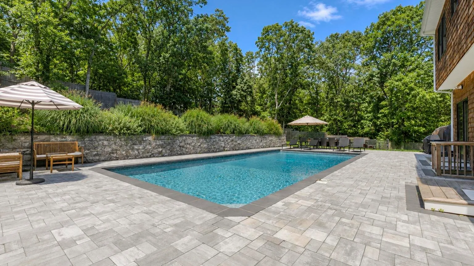 An outdoor swimming pool is surrounded by a paved patio and leafy trees.