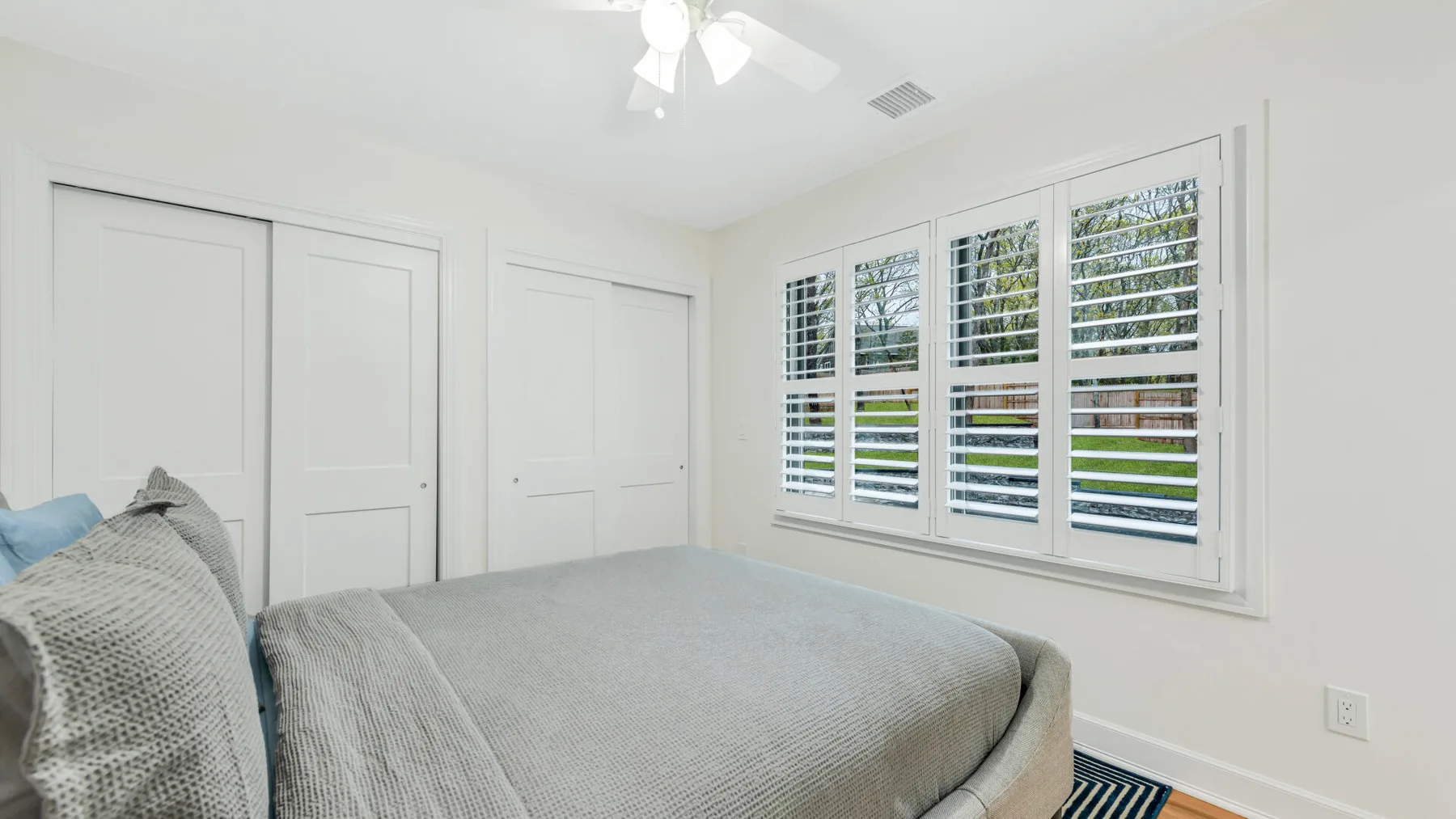 Un dormitorio luminoso con una cama tendida junto a ventanas con persianas blancas.