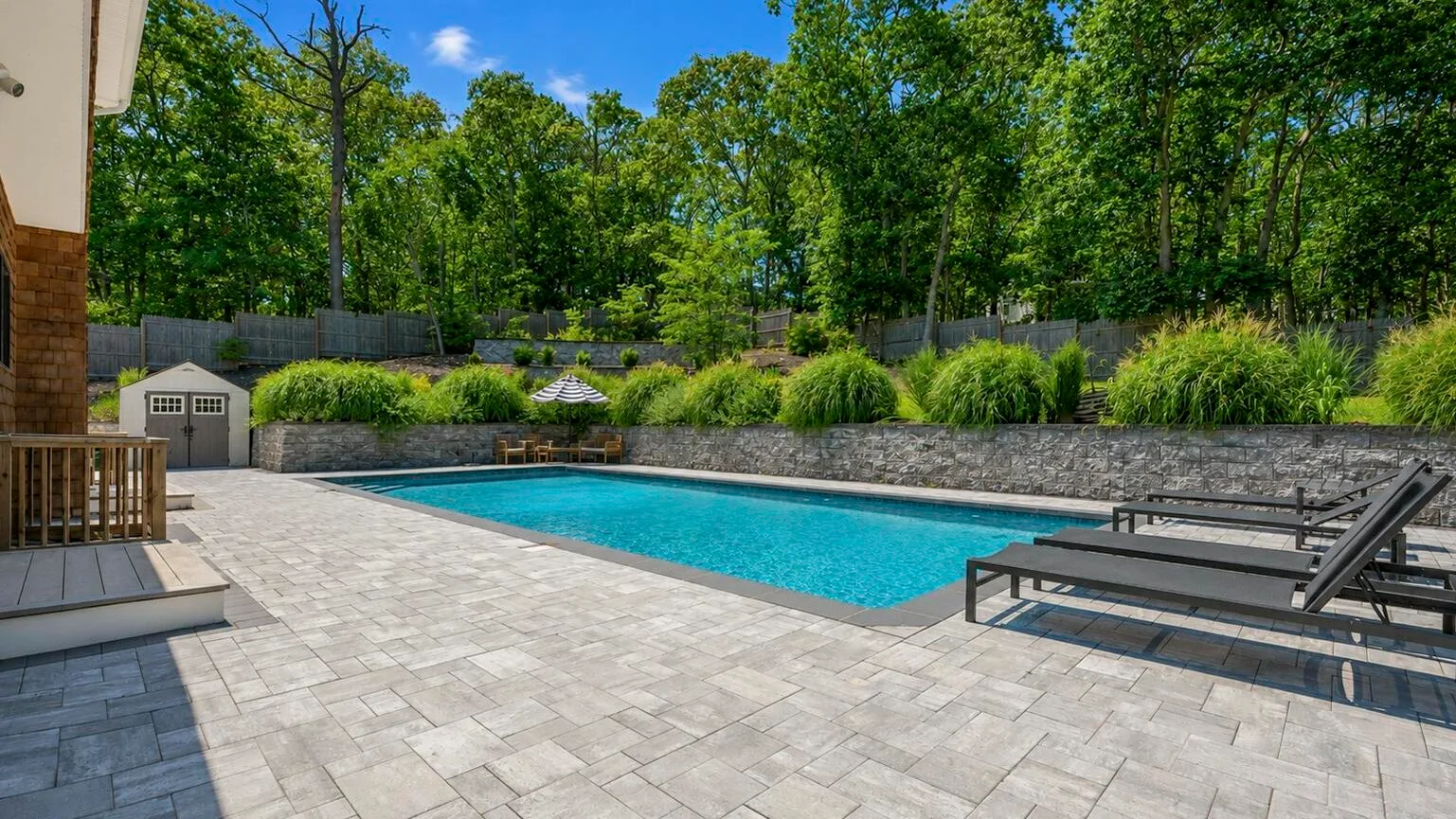 Una piscina al aire libre está junto a un patio pavimentado, con árboles al fondo.
