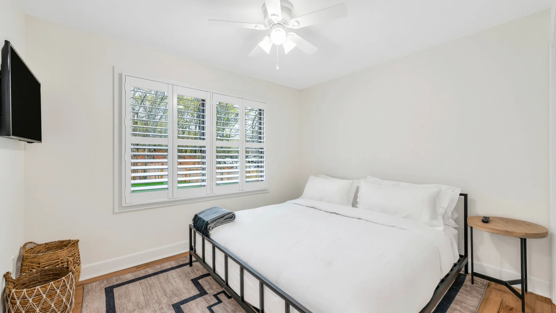 Un dormitorio luminoso con una cama blanca, una ventana con contraventanas y un ventilador de techo.